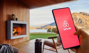 Teléfono con logo de Airbnb en pantalla roja, junto a una chimenea y paisaje. Cuidado con las estafas Airbnb.
