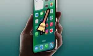 Mano sujetando un iPhone con iconos de apps y una escoba, sugiriendo la acción de borrar caché del iPad.