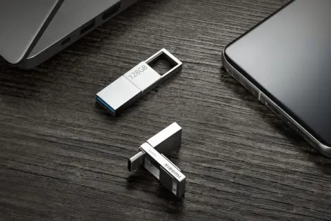 Dos unidades USB plateadas, una de 128GB y otra con logo Xiaomi, junto a un portátil y un smartphone. Perfectas para tus aplicaciones portables.
