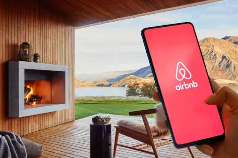 Teléfono con logo de Airbnb en pantalla roja, junto a una chimenea y paisaje. Cuidado con las estafas Airbnb.