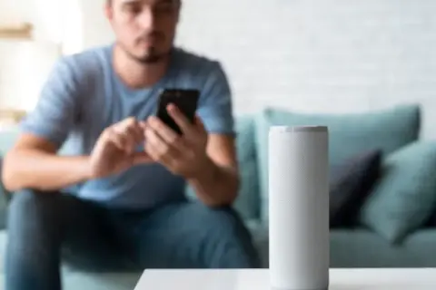 Hombre en sofá usa móvil mientras altavoz inteligente espera órdenes en modo super alexa.