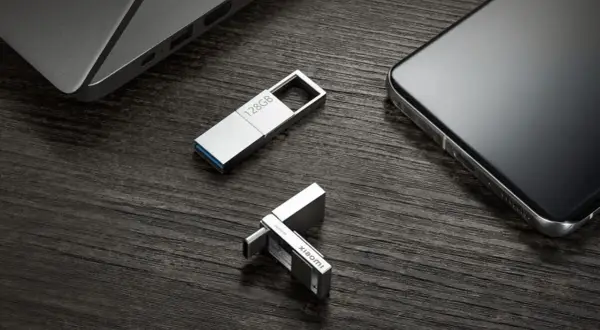 Dos unidades USB plateadas, una de 128GB y otra con logo Xiaomi, junto a un portátil y un smartphone. Perfectas para tus aplicaciones portables.