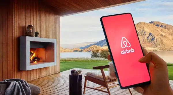 Teléfono con logo de Airbnb en pantalla roja, junto a una chimenea y paisaje. Cuidado con las estafas Airbnb.