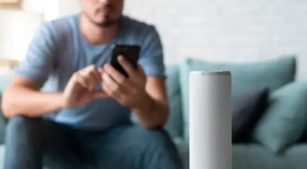 Hombre en sofá usa móvil mientras altavoz inteligente espera órdenes en modo super alexa.