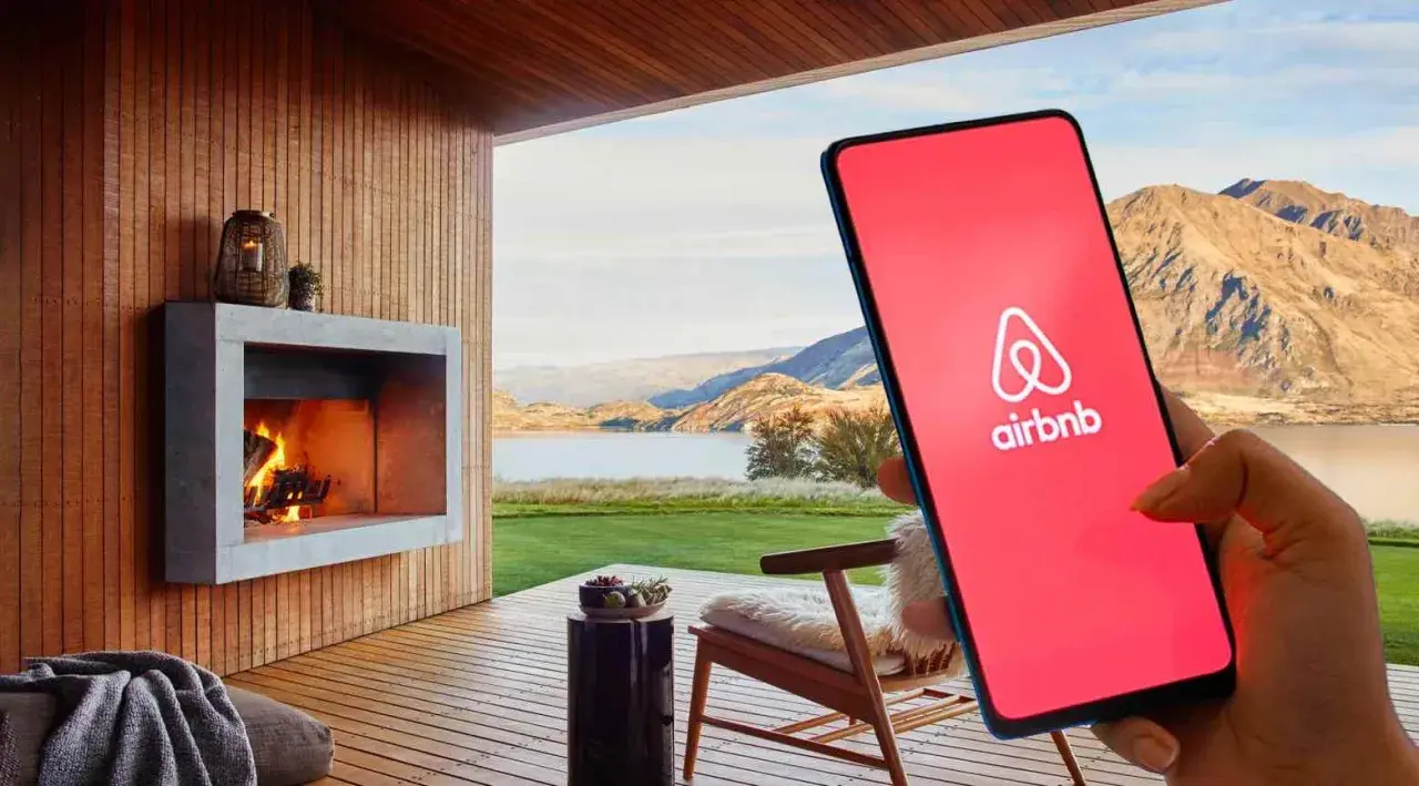 Teléfono con logo de Airbnb en pantalla roja, junto a una chimenea y paisaje. Cuidado con las estafas Airbnb.