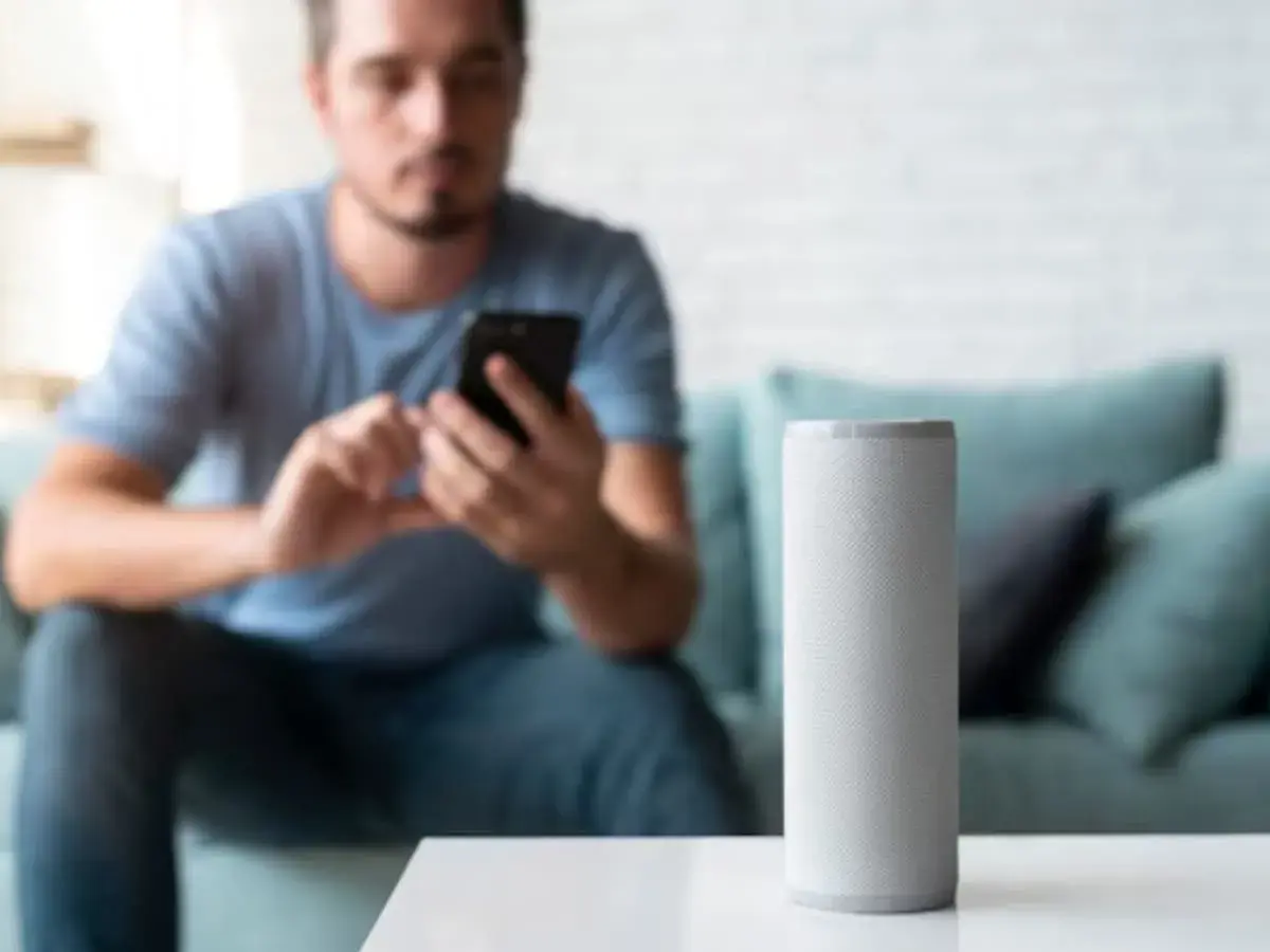 Hombre en sofá usa móvil mientras altavoz inteligente espera órdenes en modo super alexa.