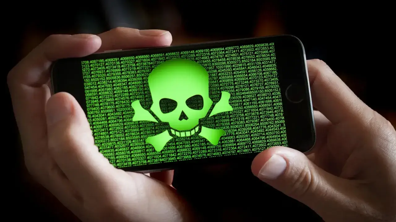 Un tel&eacute;fono muestra un cr&aacute;neo verde y huesos cruzados, s&iacute;mbolo de un virus m&oacute;vil, sobre un fondo de c&oacute;digo verde.