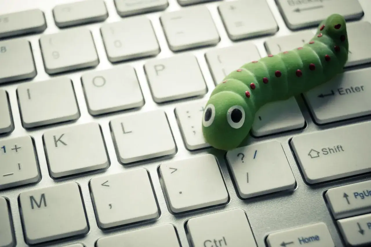 Un gusano informático verde, con puntos rojos y ojos saltones, se arrastra sobre un teclado.