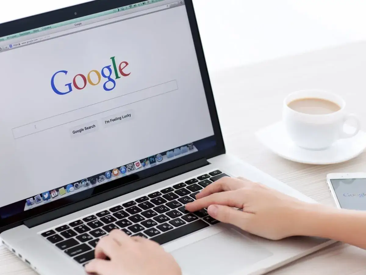 Manos tecleando en un port&aacute;til con la p&aacute;gina de Google abierta, buscando trucos de Google. Una taza de caf&eacute; y un m&oacute;vil completan la escena.
